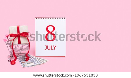A gift box in a shopping trolley, dollars and a calendar with the date of 8 july on a pink background.