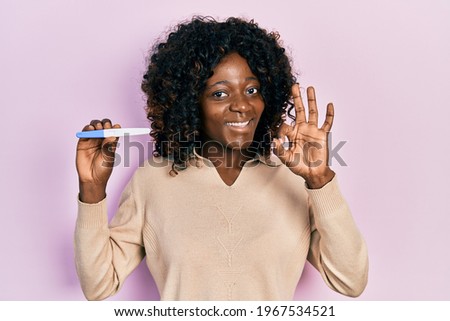 Young african american woman holding pregnancy test result doing ok sign with fingers, smiling friendly gesturing excellent symbol 
