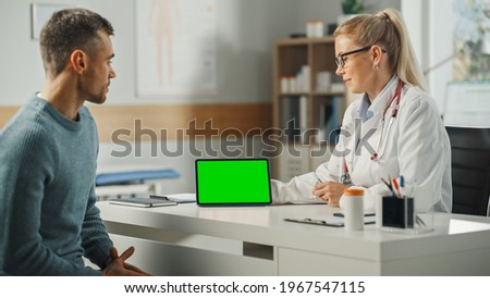 Female Physician Showing Tablet Computer with Green Screen Display to Young Male Patient During Consultation in a Health Clinic. Family Doctor Sitting Behind a Desk in Hospital Office.