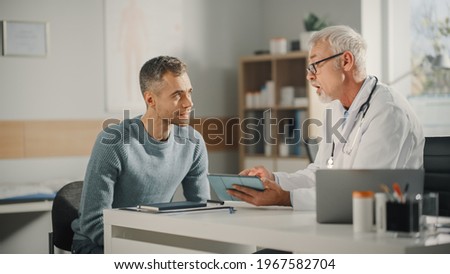 Experienced Middle Aged Family Doctor Showing Analysis Results on Tablet Computer to Male Patient During Consultation in a Health Clinic. Physician Sitting Behind a Desk in Hospital Office. Royalty-Free Stock Photo #1967582704