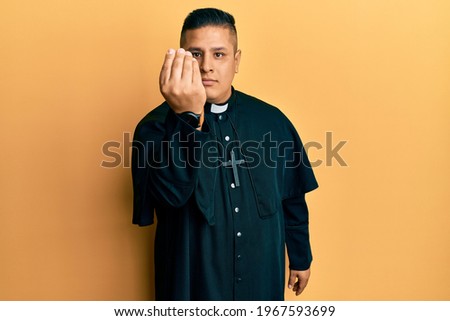 Young latin priest man standing over yellow background doing italian gesture with hand and fingers confident expression 