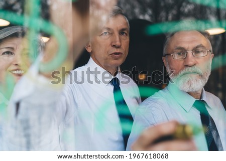 Through glass of focused male and female coworkers standing near transparent board and discussing business project together in modern workspace