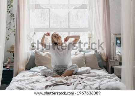 Happy young woman stretching in bed waking up in cozy bedroom with bohemian interior style. Female wearing in comfort pajamas enjoying early morning, resting alone Royalty-Free Stock Photo #1967636089