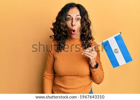 Middle age hispanic woman holding el salvador flag scared and amazed with open mouth for surprise, disbelief face 