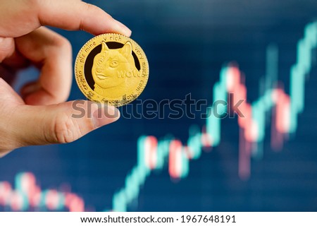 Hand holding gold Dogecoin with blurred candlestick chart in the background. DOGE is the most popular meme coin in cryptocurrency world. Royalty-Free Stock Photo #1967648191