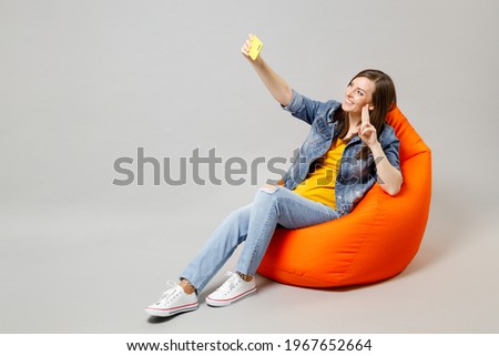 Full length young caucasian woman in casual denim jacket yellow t-shirt sitting in bean bag chair do selfie shot on mobile cell phone show victory gesture isolated on grey background studio portrait.