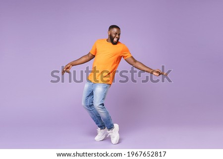 Full length of excited funny young african american man 20s in basic casual orange t-shirt dancing standing on toes pointing index fingers aside isolated on pastel violet background studio portrait Royalty-Free Stock Photo #1967652817