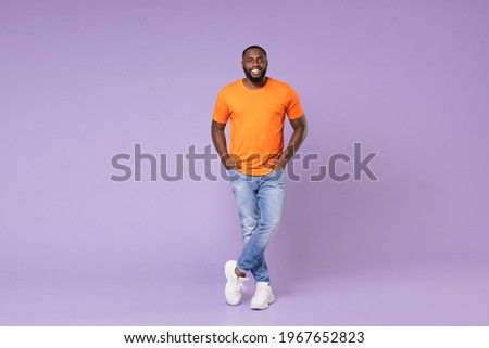 Full length side view of smiling young african american man 20s wearing basic casual orange t-shirt holding hands in pockets looking camera isolated on pastel violet colour background studio portrait