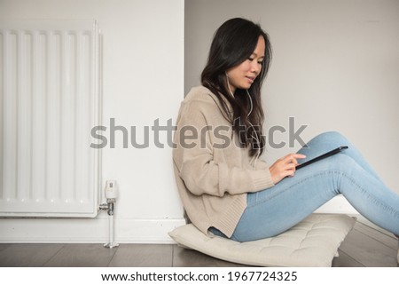 A young Asian woman sitting on the floor beside a radiator adjusts the temperature on her mobile phone as she listens to music on her earphones and smiles in the living room of a house in Edinburgh