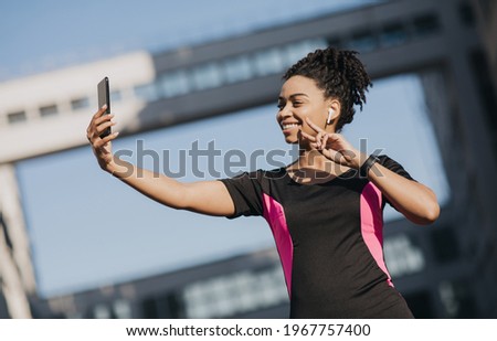 Modern sports blog, fitness model and workout with gadgets. Happy young african american woman in headphones with fitness tracker makes selfie on smartphone, on city building and sky background