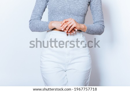 Close-up of folded arms of woman dressed in white high-waisted jeans and gray long-sleeve crop top, standing against white studio background.