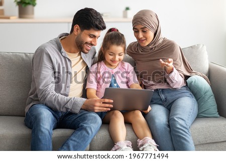 Happy muslim parents teaching daughter using laptop, sitting together on sofa at home. Family surfing internet or having video chat with someone. Arab man, child and woman in hijab staying home