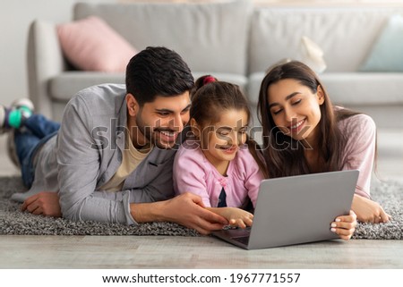 Family weekend. Cheerful arab parents teaching their little daughter using laptop, spending time at home together. Father, mother and girl searching cartoon in internet, lying on the floor carpet