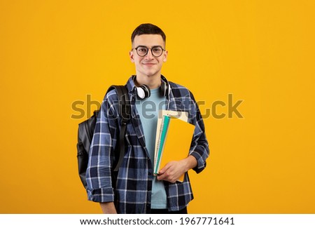 Education concept. Positive young man with backpack, headphones and notebooks posing on orange studio background. Cool millennial student smiling and looking at camera Royalty-Free Stock Photo #1967771641