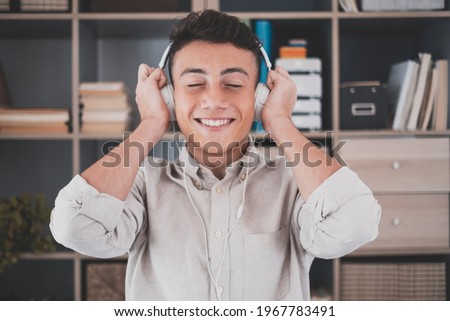 Tranquil carefree young teenager man at home, wearing modern wireless headphones, listening to favorite classic music online, feeling peaceful mindful alone in living room.