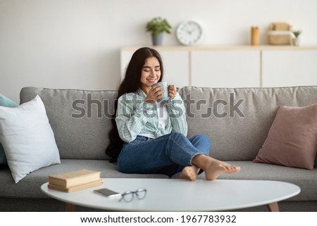 Full length of young Indian woman having coffee break on comfy sofa at home. Beautiful millennial lady relaxing on couch, enjoying lazy weekend morning with hot beverage in living room