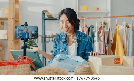 Young Asia lady fashion designer using mobile phone receiving purchase order and showing clothes recording video live streaming online at shop. Small business owner, online market delivery concept. Royalty-Free Stock Photo #1967798545