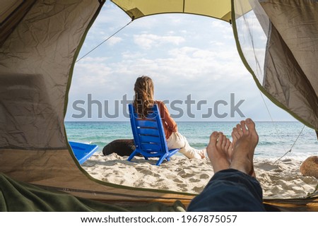 Camping on the beach. Unrecognizable man inside the tend and rear view of woman sitting on the beach shore with a dog.