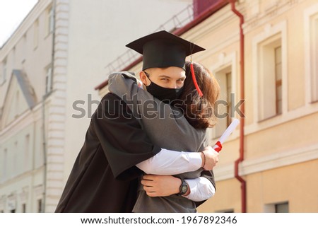Mother hugs her son studentin face mask in graduation gown and a square cap after graduation ceremony. Class 2021
