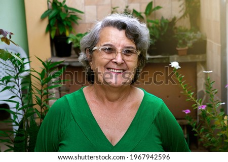Elderly, senior, mature woman in home garden smiling standing looking at camera front view. Pictured from the home garden, natural light. Sun light. Healthy living in old age. Beautiful garden.