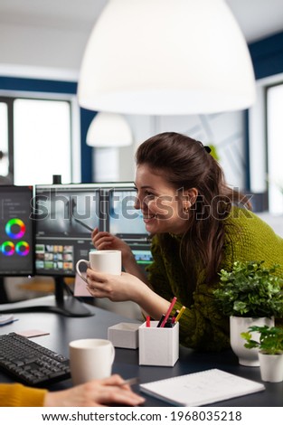 Happy videographer editing video project drinking coffee using post production software working in digital multimedia company with dual monitors setup. Retoucher processing audio film montage on pc
