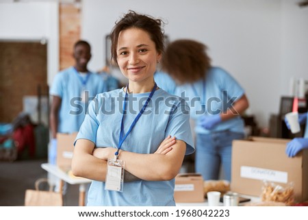 Portrait of happy young woman, volunteer in blue uniform smiling at camera while standing with arms crossed indoors. Team sorting, packing items in the background Royalty-Free Stock Photo #1968042223