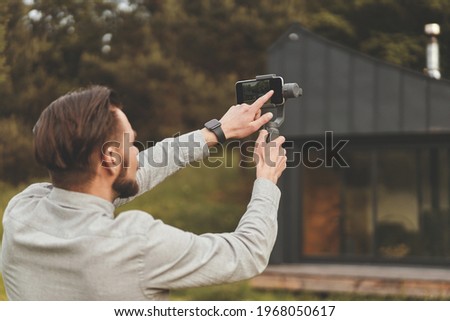 Back view of millennial man recording video at smartphone with tripod. Bearded man with smartwatch standing outside in front of modern country house, holding tripod and recording video