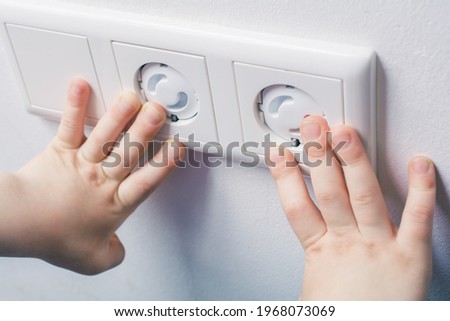 Two Child Hands Touching A Wall Socket With Safety Plugs - Prevent Child Hazard Concept Royalty-Free Stock Photo #1968073069
