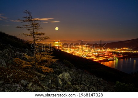 Picturesque night landscape. View from the mountain to the night city. Full moon in the sky over the city and the sea bay. Port town on the sea coast. Magadan, Magadan Region, Siberia, Far East Russia Royalty-Free Stock Photo #1968147328