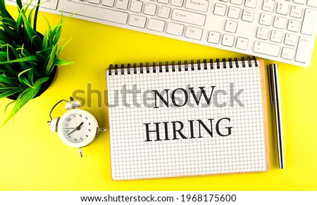 NOW HIRING text on notebook with keyboard , pen and alarm clock on yellow background