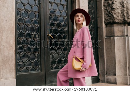 Street style, fashion conception: elegant woman wearing trendy pink suit, burgundy color hat, holding yellow bag, posing in street of city. Copy, empty space for text Royalty-Free Stock Photo #1968183091
