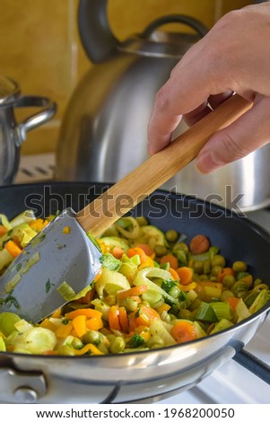 Process of cooking saute at home. Woman's hand mixed stewed vegetables in frying pan on gas stove with silicone spatula with wooden handle. Ingredients leek, carrot, zucchini, bell pepper, green peas. Royalty-Free Stock Photo #1968200050