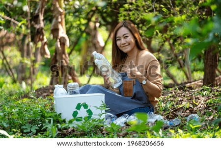 A beautiful asian woman making thumbs up hand sign while collecting garbage plastic bottles into a recycle bin in the outdoors