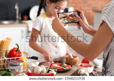 Female food photographer with mobile phone and little girl cooking pizza in kitchen