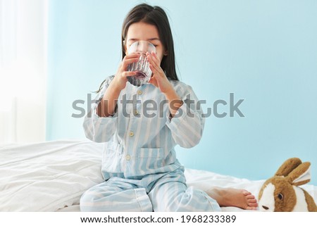 Horizontal image of a little girl drinking fresh water in the bed at home. Cute preschool kid holding glass of pure water in the morning. Healthy lifestyle concept.