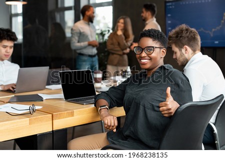 Successful Meeting. Portrait of confident smiling African American businesswoman in eyeglasses sitting at desk in office with colleagues, showing thumbs up gesture, posing and looking at camera