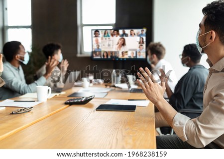 Videoconferencing Concept. Diverse group of workers in face masks making video call with colleagues, sitting at table in boardroom, waving at screen at office, talking on web with remote employees Royalty-Free Stock Photo #1968238159