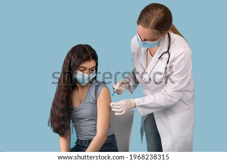 Covid-19 immunization concept. Teenage girl being vaccinated against coronavirus on blue studio background. Young doctor giving antiviral vaccine injection to adolescent patient Royalty-Free Stock Photo #1968238315