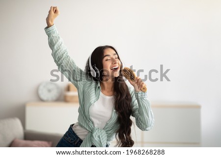 Happy Indian lady with headphones dancing to music and singing song, using hairbrush as mic at home. Young woman moving to her favorite soundtrack, pretending to be rock star Royalty-Free Stock Photo #1968268288