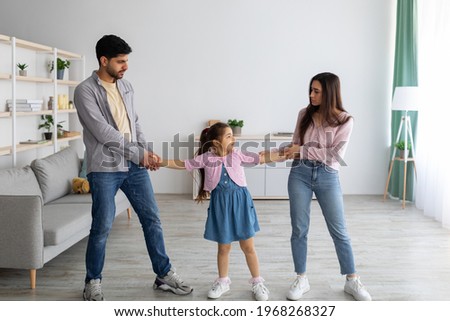 Divorce concept. Arab parents fighting over their child, mad man and woman quarrelling, pulling daughter's hands in different directions, standing in living room interior Royalty-Free Stock Photo #1968268327