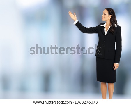 Full body smiling woman in black confident suit, showing advertising at copy space. Business ad concept. Blurred modern office interior background. Brunette businesswoman, profile side.