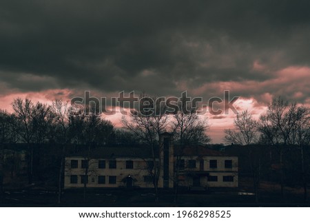 An old building in the village, storm clouds over the roof. Red sunset against the background of trees and buildings. Empty streets and abandoned houses. Evening Royalty-Free Stock Photo #1968298525