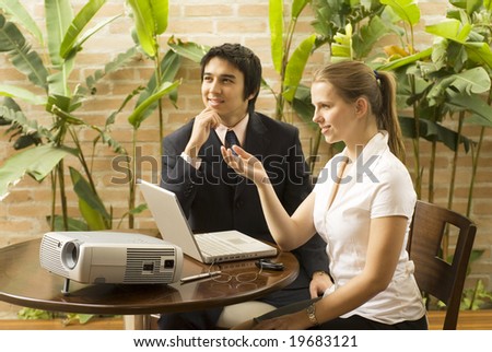 Man and woman with a projector and a laptop. They are seated and smiling . Horizontally framed photo.