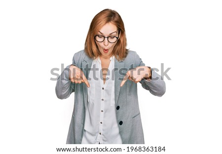Young caucasian woman wearing business style and glasses pointing down with fingers showing advertisement, surprised face and open mouth 