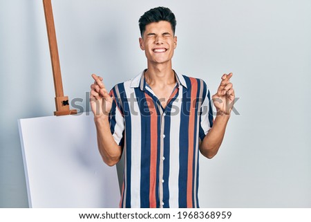 Young hispanic man standing close to empty canvas gesturing finger crossed smiling with hope and eyes closed. luck and superstitious concept. 