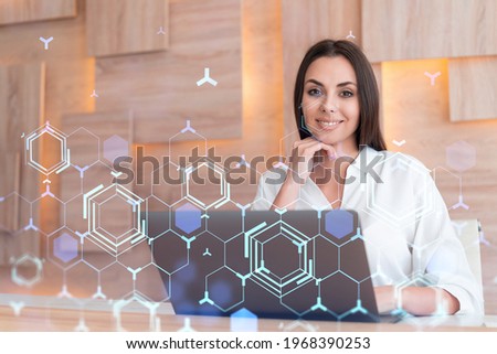 Attractive businesswoman in white shirt at workplace working with laptop to optimize development by implying new technologies in business process. Hi tech hologram over office background
