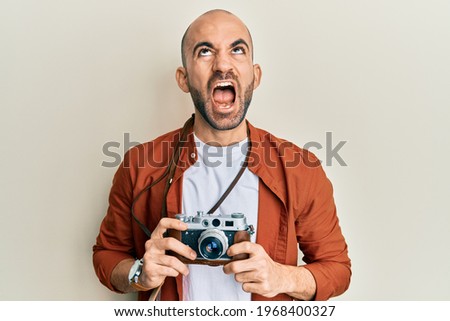 Young hispanic man holding vintage camera angry and mad screaming frustrated and furious, shouting with anger looking up. 