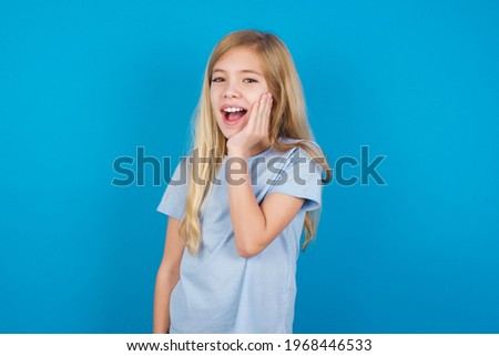Shocked, astonished beautiful Caucasian little girl wearing blue T-shirt over blue background looking surprised in full disbelief wide open mouth with hand near face. Positive emotion facial expressio