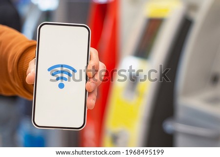 Online payments. Phone display with wi-fi icon against the backdrop of a ATM cash machine. Man holds a smartphone in his hand close-up