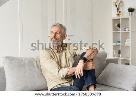 Relaxed mature older thoughtful tattooed hipster man relaxing sitting on sofa and thinking at home. Pensive calm stylish mid aged single bearded guy resting on couch at home looking away, reflecting. Royalty-Free Stock Photo #1968509698